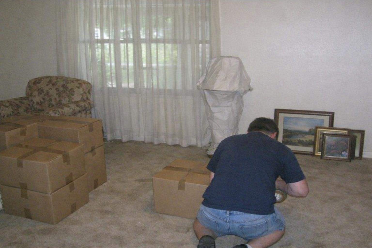 An older living room being cleared, with taped boxes of books, a wrapped lamp and a floral armchair